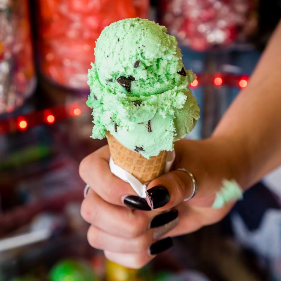 A hand holding a cone of mint chocolate chip ice cream in front of the candy shelves