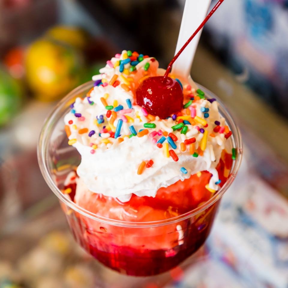 A strawberry sundae in a clear cup topped with whipped cream, rainbow sprinkles and a cherry